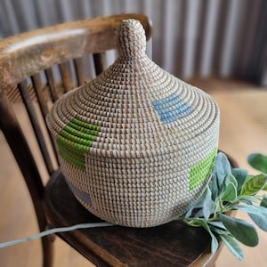 May include: A woven basket with a lid, featuring a white base with green and blue geometric patterns. The basket is sitting on a wooden chair.