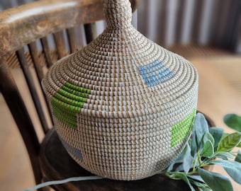 May include: A woven basket with a lid, featuring a white base with green and blue geometric patterns. The basket is sitting on a wooden chair.