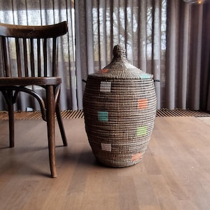 Puede incluir: Una cesta ovalada tejida con tapa, con un patrón marrón y negro con acentos cuadrados turquesa, naranja y verde. La cesta está sobre un suelo de madera. A la izquierda hay una silla de madera. La cesta mide unos 50 cm de altura.