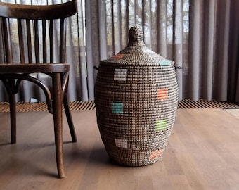 May include: A woven basket with a lid, featuring a brown and black pattern with colorful square accents in teal, orange, and green. The basket is oval-shaped and sits on a wooden floor. A wooden chair is to the left.