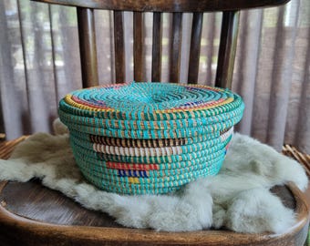 May include: A round, woven basket with a lid. The basket is turquoise with bands of yellow, red, and white. It sits on a wooden chair. The basket is likely handmade and suitable for storage or decoration.
