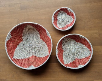May include: Three woven baskets in varying sizes, featuring a red and white geometric design. The baskets are arranged on a wooden surface, showcasing their circular shape and textured weave. The largest basket is approximately 12 inches in diameter.