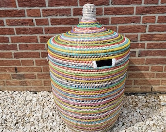 May include: A large, cylindrical woven basket with a lid. The basket is adorned with horizontal bands of various colors, including yellow, blue, green, and pink. The lid has a conical shape and is topped with a woven knob. The basket has a rectangular opening.