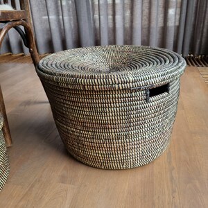 May include: A round woven basket with a lid. The basket is made of natural fibers in shades of black, tan, and cream. It has a circular lid and a rectangular handle cut-out. The basket is sitting on a wooden floor.
