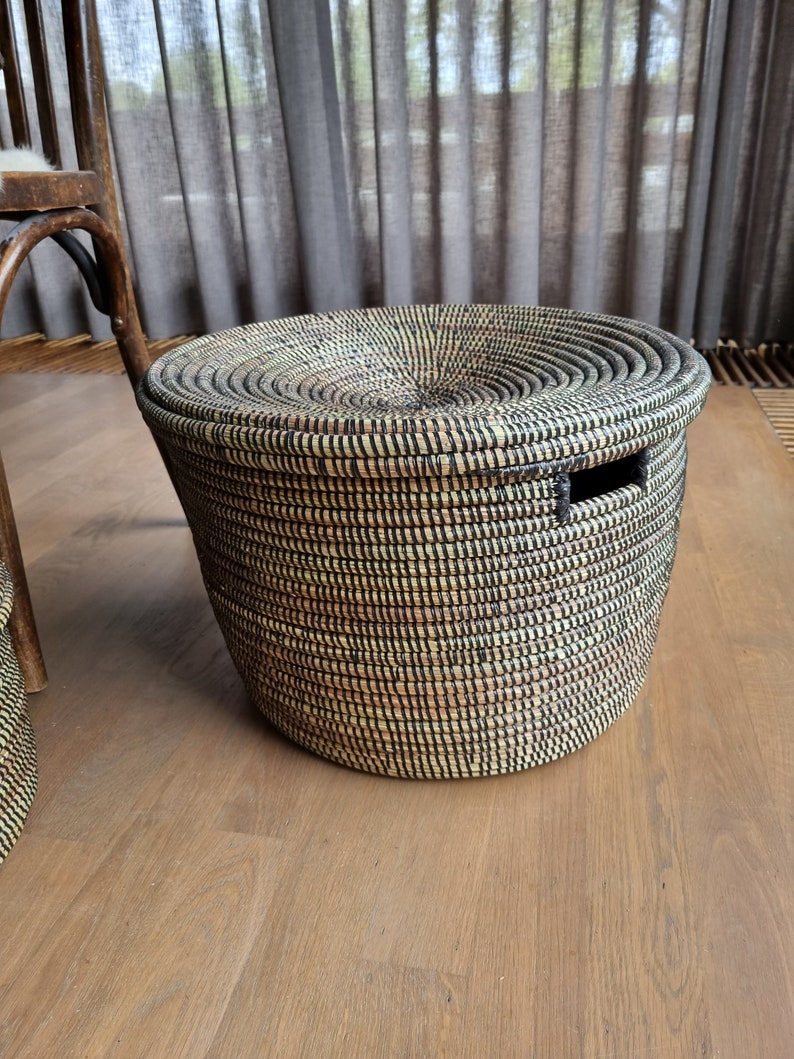 May include: A round woven basket with a lid. The basket is made of natural fibers in shades of black, tan, and cream. It has a circular lid and a rectangular handle cut-out. The basket is sitting on a wooden floor.