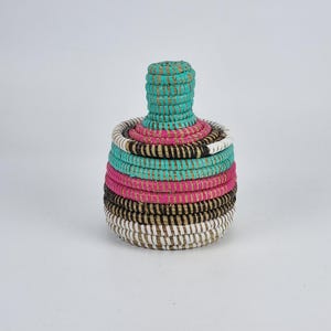 Op de afbeelding: Een kleine, handgemaakte mand met een deksel. De mand is gemaakt van geweven natuurlijke vezels en heeft banden in turquoise, roze, zwart en wit. Het deksel is cilindrisch en turquoise. De mand wordt waarschijnlijk gebruikt voor opslag of decoratie.
