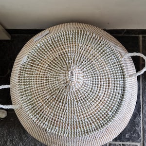 May include: A round, woven basket with a lid, featuring a concentric pattern of white and natural tan fibers. The basket includes two white braided handles on opposite sides and a small, rectangular white tag attached to the top surface.
