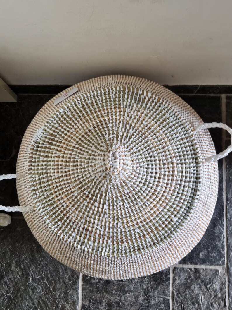 May include: A round, woven basket with a lid, featuring a concentric pattern of white and natural tan fibers. The basket includes two white braided handles on opposite sides and a small, rectangular white tag attached to the top surface.