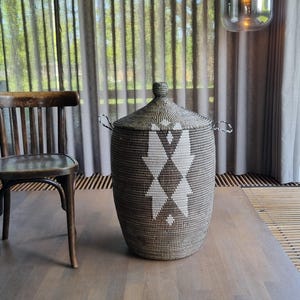 Op de afbeelding: Een cilindrische geweven opbergmand met een deksel. De mand heeft een geometrisch ruitpatroon in wit en bruin. Hij heeft twee handvatten en een knop op het deksel. De mand staat op een houten oppervlak.