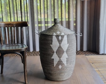 May include: A woven storage basket with a lid. The basket is cylindrical with a geometric diamond pattern in white and brown. It has two handles and a knob on the lid. The basket is on a wooden surface.