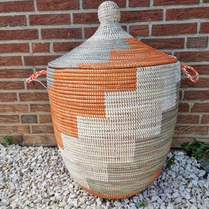 Puede incluir: Una cesta de almacenamiento grande tejida a mano con tapa cónica y remate esférico. La cesta presenta un patrón geométrico en tonos naranja vibrante, crema y gris, con dos asas laterales trenzadas en color naranja y blanco.