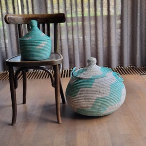 Op de afbeelding: Twee geweven opbergmanden met deksels. Eén mand staat op een houten stoel, de andere op de vloer. De manden zijn teal, grijs en naturel van kleur, met een ronde vorm. De deksels hebben een knop bovenop.