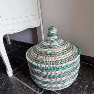 May include: A round woven basket with a lid. The basket is made of natural fibers and features a pattern of green, white, and tan stripes. The lid has a conical handle. The basket is sitting on a dark tiled floor.