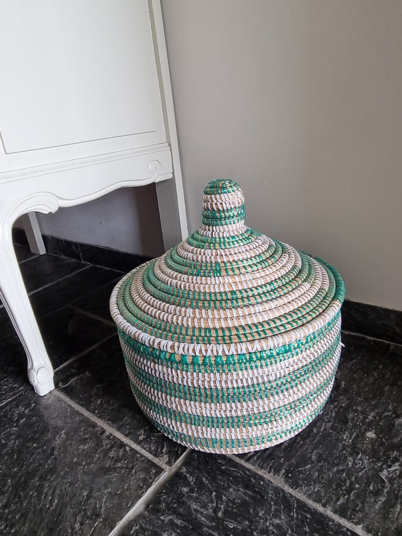 May include: A round woven basket with a lid. The basket is made of natural fibers and features a pattern of green, white, and tan stripes. The lid has a conical handle. The basket is sitting on a dark tiled floor.