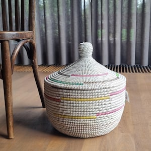 May include: A round, woven storage basket with a matching conical lid and a small knob handle. The basket is primarily white with horizontal bands of yellow, pink, and green accents woven into the structure. It features a textured, coiled design.