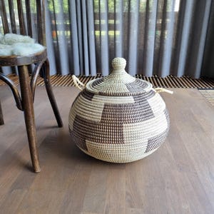 May include: A spherical basket with a lid, featuring a woven pattern in brown and beige. The basket has a small knob and two handles. It rests on a wooden floor.