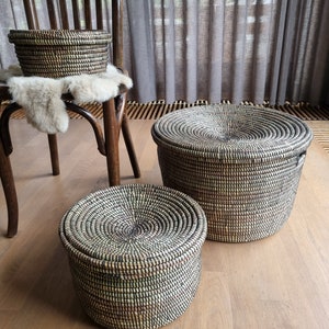 May include: Three woven storage baskets with lids in different sizes. The baskets are black and beige, with a circular pattern on the lids. One basket is on a wooden chair, the others on a wooden floor.