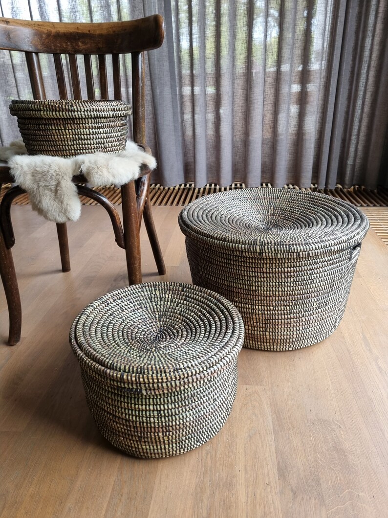 May include: Three woven storage baskets with lids in varying sizes. The baskets are black and beige, with a circular design on the lids. One basket sits on a wooden chair, and the others are on a wooden floor.