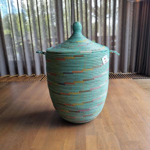 Op de afbeelding: Een hoge, cilindrische gevlochten mand in een licht mintgroene kleur met een bijpassend conisch deksel en twee handvatten aan de zijkant. De mand heeft horizontale, gestreepte accentlijnen in oranje, geel, roze en donkerblauw, met een klein wit label.