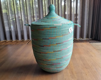 May include: A tall, cylindrical woven basket in a light mint green color with a matching conical lid and two side handles. The basket features horizontal, dashed accent lines in orange, yellow, pink, and dark blue, with a small white rectangular tag.