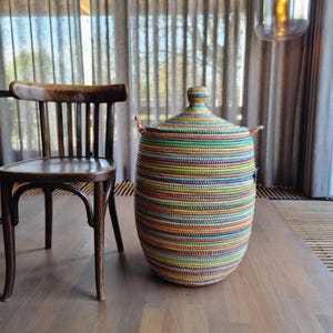 Op de afbeelding: Een hoge, cilindervormige opbergmand met een conisch deksel en twee kleine handvatten aan de zijkant. De mand is gemaakt van strak geweven, veelkleurig materiaal met horizontale strepen in de kleuren groen, blauw, geel, oranje en rood.