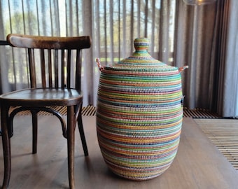 May include: A tall, cylindrical storage basket with a conical lid and two small side handles. The basket is constructed from tightly coiled, multi-colored material featuring horizontal stripes in shades of green, blue, yellow, orange, and red.