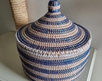 May include: A round woven basket with a lid. The basket is made of natural-colored fibers, with blue and white stripes. The lid has a knob on top. A smaller woven item is in the background.