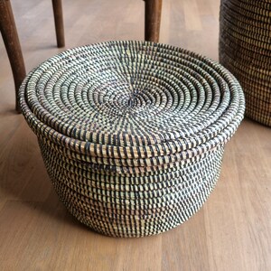 May include: A round, lidded basket with a woven design in natural and black tones. The basket is made of tightly woven fibers, creating a textured surface. The lid fits snugly on top. A second basket is partially visible in the background.