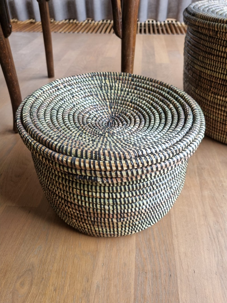 May include: A round, lidded basket with a woven design in natural and black tones. The basket is made of tightly woven fibers, creating a textured surface. The lid fits snugly on top. A second basket is partially visible in the background.