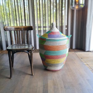 Op de afbeelding: Een grote, kleurrijke geweven mand met een deksel, met een levendige mix van kleuren, waaronder blauw, groen, geel en roze. De mand staat op een houten oppervlak naast een houten stoel. De mand heeft twee groene handvatten.