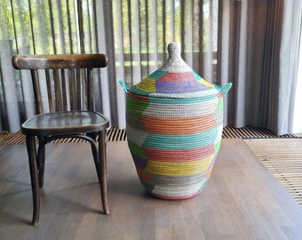 May include: A large, colorful woven basket with a lid, featuring a vibrant mix of colors including blue, green, yellow, and pink. The basket is placed on a wooden surface next to a wooden chair. The basket has two green handles.