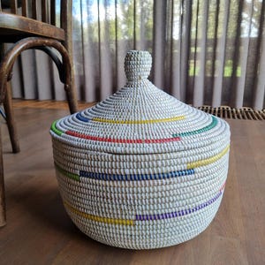 May include: A round, white woven basket with a matching conical lid featuring a small knob handle. The basket is decorated with horizontal, intermittent stripes in vibrant colors including red, blue, yellow, green, and purple throughout the body.