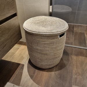 May include: A white and brown woven storage basket with a lid. The basket is round and has a woven pattern. It is sitting on a wooden floor.