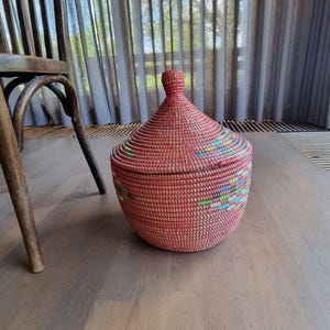 May include: A red, lidded basket with a conical top. The basket has bands of colourful woven threads around the base. It sits on a wooden floor next to a wooden chair. The basket is likely hand-woven.