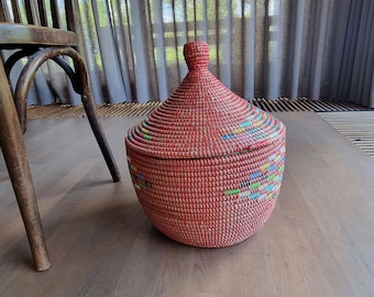 May include: A round, lidded basket with a conical top. The basket is primarily red with colorful bands of woven threads around the base. The basket sits on a wooden floor next to a wooden chair.