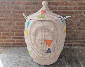 May include: A large, woven basket with a lid. The basket is beige with colorful triangle patterns in orange, blue, green, and pink. It has two handles and a knob on the lid. The basket is sitting on a stone surface in front of a brick wall.