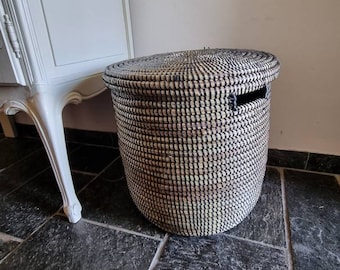 May include: A round, lidded storage basket featuring a woven pattern of alternating black and natural tan fibers. The basket has a cylindrical shape with a flat, circular lid and integrated side handles cut into the upper rim of the structure.