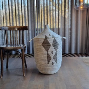 Op de afbeelding: Een geweven opbergmand met een deksel, met een ruitpatroon in grijstinten en bruin. De mand heeft twee kleine handvatten en staat op een houten vloer. Links staat een houten stoel.