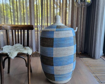 May include: A large, woven storage basket with a lid. The basket features a checkerboard pattern in shades of blue, gray, and brown. It has two white rope handles and sits on a wooden surface. The basket is approximately 24 inches tall.