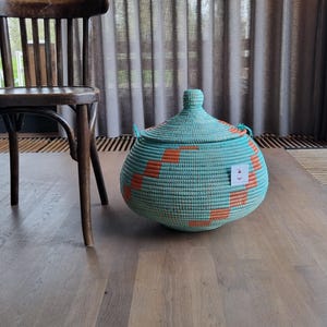 May include: A teal woven basket with an orange geometric pattern and a lid. The basket is round with small handles. A wooden chair is in the background. The basket is approximately 30 cm in diameter.