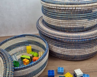 May include: Three round woven baskets with blue and white stripes. The baskets are stacked on top of each other. The top basket is open and has a lid. The baskets are filled with colorful plastic building blocks.