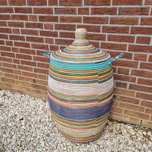 Puede incluir: Una cesta grande y redonda tejida con tapa. La cesta está adornada con bandas horizontales de varios colores, incluyendo turquesa, amarillo, rosa y azul. Tiene dos asas pequeñas y un pomo en la tapa. El fondo es una pared de ladrillos rojos.