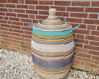 May include: A large, round woven basket with a lid. The basket is adorned with horizontal bands of various colors, including teal, yellow, pink, and blue. It has two small handles and a knob on the lid. The background features a red brick wall.