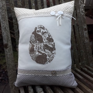 Può includere: Un cuscino bianco con un disegno ricamato marrone di un cerchio con conigli e fiori al suo interno. Il cuscino ha un bordo in pizzo bianco e un tessuto a pois marroni sul retro.