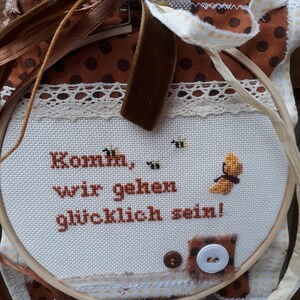 Könnte beinhalten: Ein weißer Stickrahmen mit dem Text "Komm, wir gehen glücklich sein!" in braunem Garn. Der Rahmen ist mit braunem und weißem Stoff, Spitze und einem braunen Band verziert.