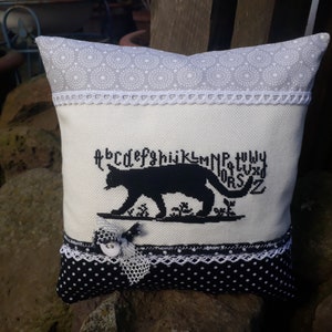 Op de afbeelding: Een wit kussen met een zwart katten silhouet geborduurd op de voorkant. De kat loopt over de letters van het alfabet. Het kussen heeft een witte kantversiering en een zwart-witte stippenstof aan de achterkant.