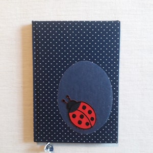 Pode incluir: Um caderno azul marinho com bolinhas brancas e um oval azul escuro. Uma joaninha vermelha e preta está no oval. Um pequeno pingente transparente pende da parte inferior do caderno.