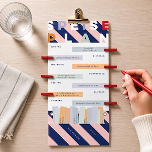 Könnte beinhalten: Ein farbenfroher Wochenessensplaner mit dem Text "SPEISE PLAN" in fetter Schrift. Der Planer hat ein gestreiftes Design in Rosa, Blau und Weiß, mit roten Clips, die Essenskarten halten. Ein Glas Wasser und ein Stift sind ebenfalls sichtbar.