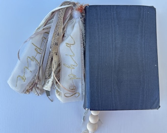 May include: A black journal with a gold tassel bookmark that says "everyday" and "so-holiday". The bookmark is made of fabric and has a wooden bead chain.
