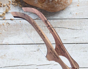 May include: Two wooden bread slicers with engraved text, "Slice of Love" and "Slice of Happiness," alongside names and dates. A loaf of bread sits in the background on a white wooden surface. The slicers have a curved design and a thin blade.
